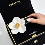 A close-up view of elegant, well-manicured hands carefully opening a pristine white luxury gift box adorned with a camellia flower. Inside, a soft black velvet dust bag is being revealed, partially showing the corner of a black Chanel Classic Flap Wallet crafted from textured caviar leather, with a gleam of its gold CC logo. The setting is a clean, minimalist surface with soft, diffused lighting, emphasizing luxury and anticipation. Perfect anatomy, well-formed hands, proper finger count, correct proportions, natural pose. Professional photography, high quality, fully clothed, appropriate attire, safe for work, appropriate content, modest, family-friendly.